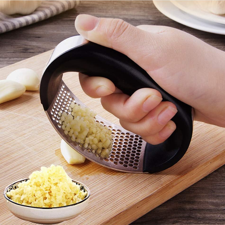 Hand pressing a black-handled rocking garlic press over garlic on a wooden cutting board.