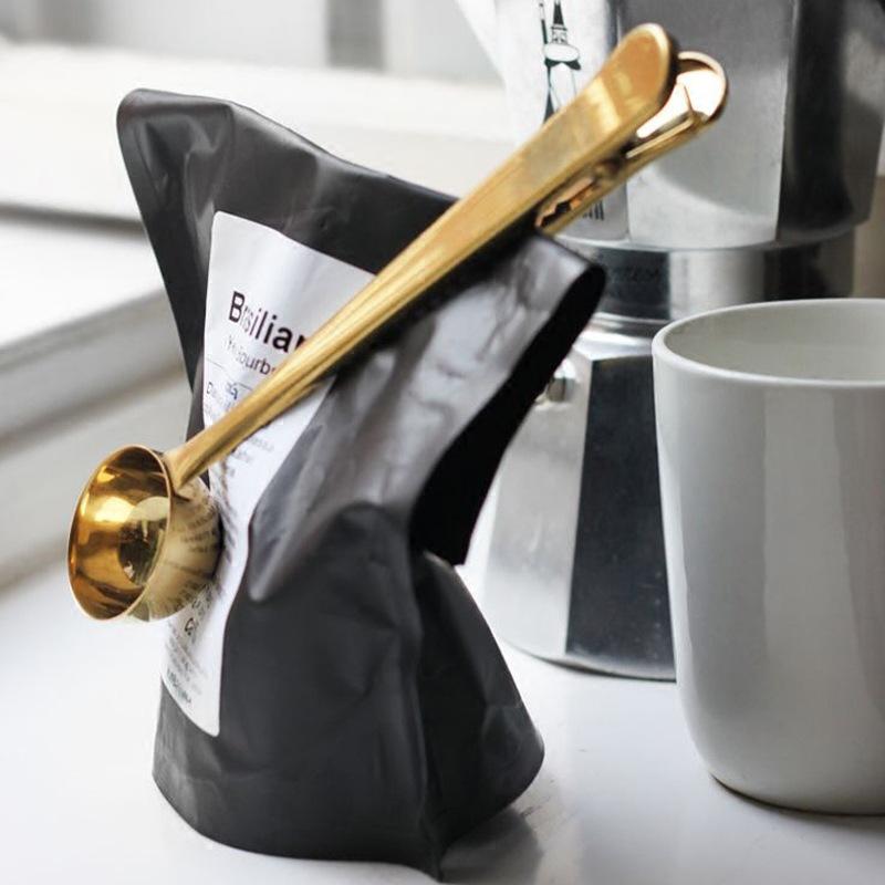 Gold coffee scoop clipped across a folded coffee bag beside a white mug and moka pot.