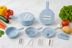 A set of light blue plastic kitchen utensils and vegetable choppers arranged on a white marble surface, surrounded by fresh vegetables including lettuce, tomato, bell pepper, carrots, cucumber, garlic, and potato.