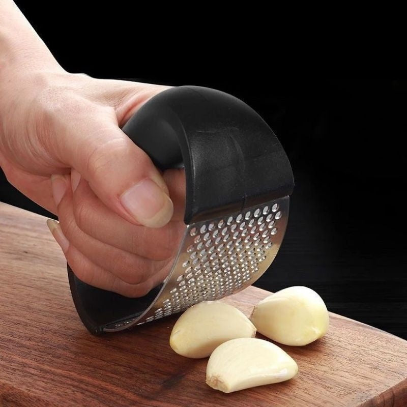 Hand holding a rocking garlic press beside peeled garlic cloves on a wooden board.