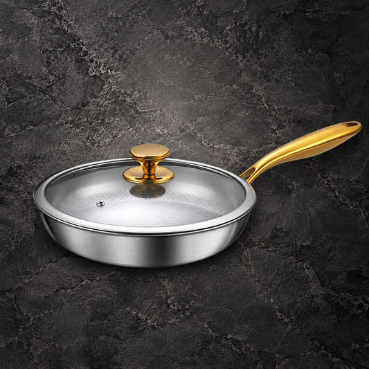 A stainless steel frying pan with a clear glass lid and polished gold-colored handle and knob, set against a dark textured stone background.