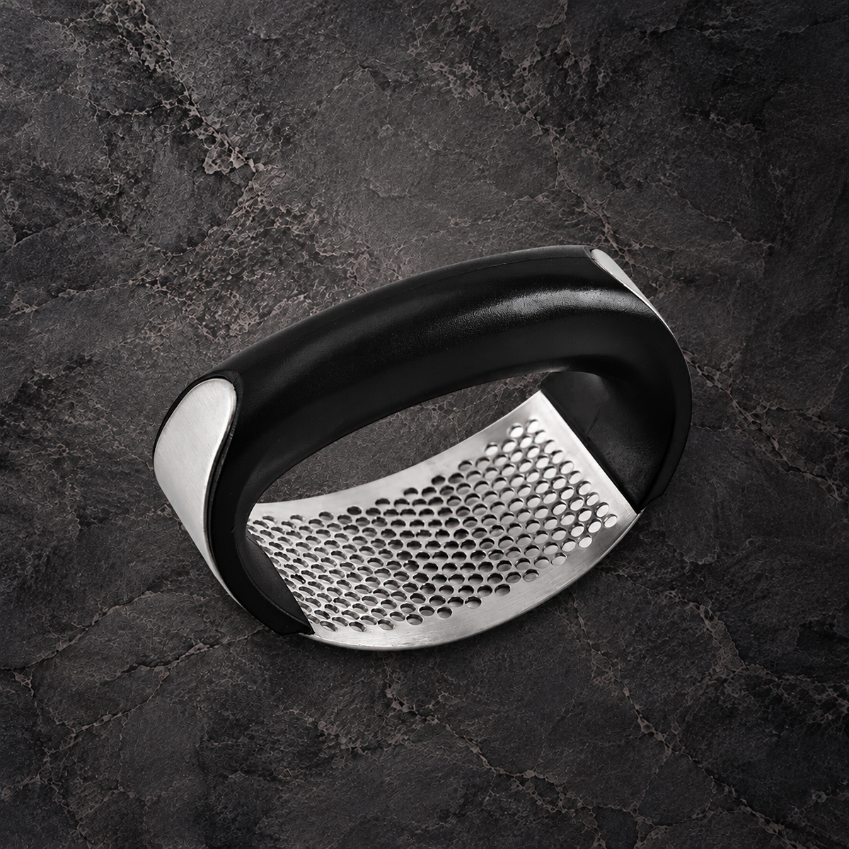 Rocking garlic press with a black curved handle and perforated stainless steel plate on dark stone.