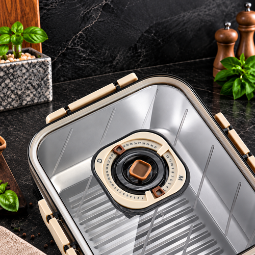 A stainless steel automatic coffee maker with beige handles and a beige dial, shown on a dark kitchen countertop with basil plants and wooden pepper mills in the background.