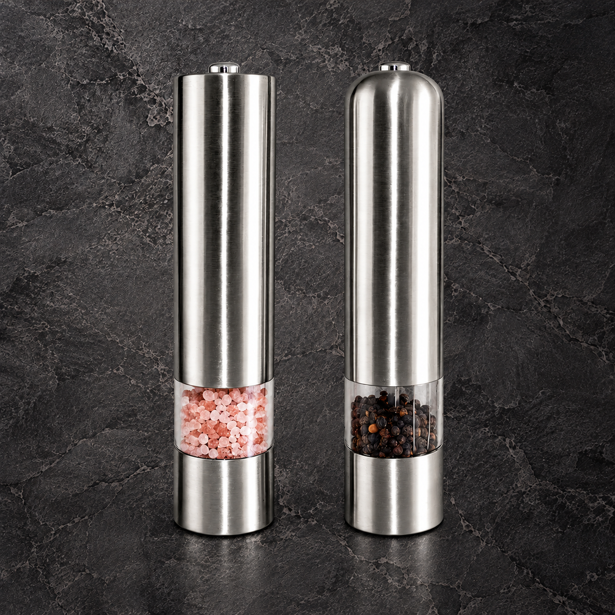 Two stainless steel electric pepper and salt mills standing side by side on a dark textured stone surface. The left mill contains pink Himalayan salt, and the right mill contains black peppercorns, both with clear windows showing the contents.