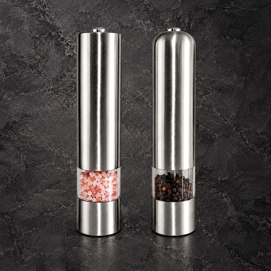 Two stainless steel electric pepper and salt mills standing side by side on a dark textured stone surface. The left mill contains pink Himalayan salt, and the right mill contains black peppercorns, both with clear windows showing the contents.