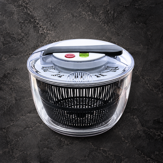 A salad spinner with a clear plastic outer bowl, black inner basket, and a black handle on top, set against a dark textured stone background.