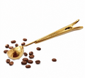 A gold-colored metal coffee scoop with a spring-loaded clip handle, shown with scattered coffee beans on a plain white background.