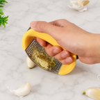 A hand using a yellow and stainless steel garlic press grater to crush a garlic bulb on a white marble surface.