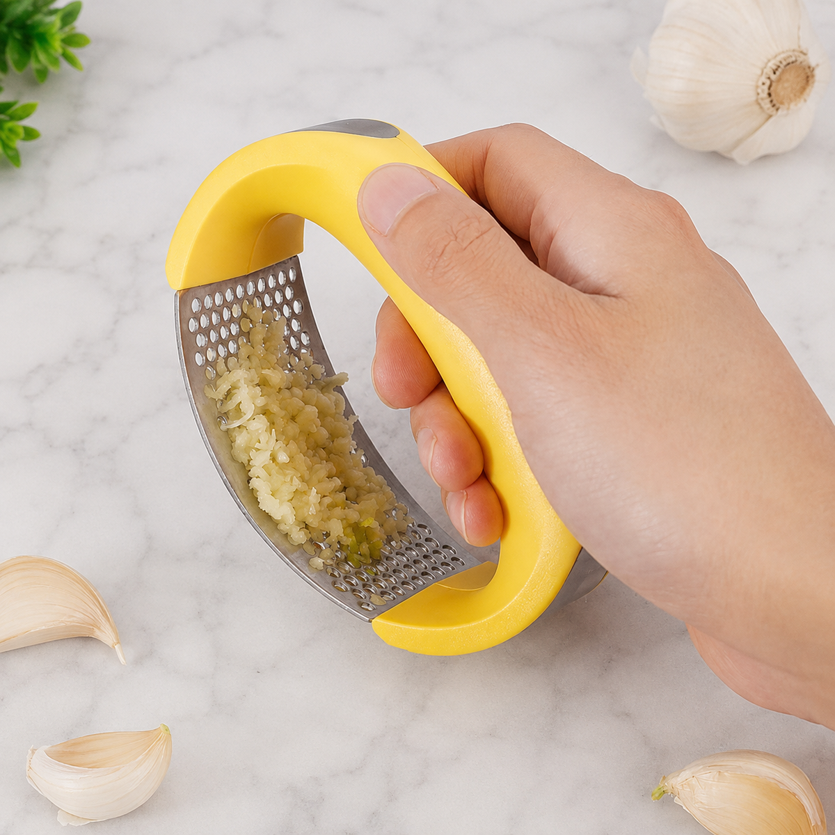 A hand holding a yellow and stainless steel garlic press grater over a white marble surface, with garlic cloves and a garlic bulb arranged around it.