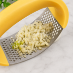 A close-up of a yellow-handled metal grater in use, with minced pale yellow food and a small piece of green peel being scraped onto a white marble surface.