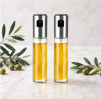 Two clear glass oil bottles with brushed silver metal caps, filled with golden yellow oil, standing on a light marble surface with olive branches in the background.