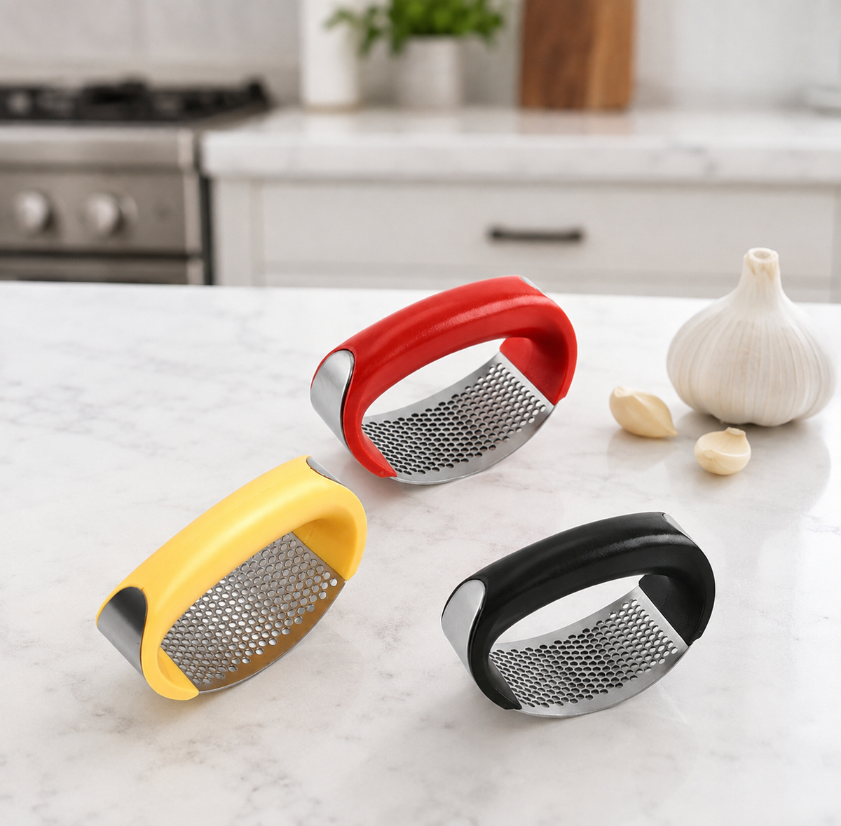 Three garlic press tools in red, yellow, and black are arranged on a white marble countertop with a garlic bulb and cloves and a blurred kitchen background.