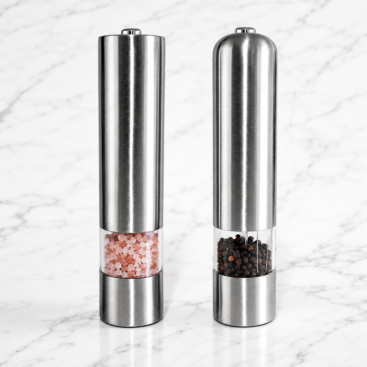 Two stainless steel pepper and salt mills standing side by side on a white marble surface. The left mill contains pink Himalayan salt, and the right mill contains black peppercorns, both with clear windows showing the contents.